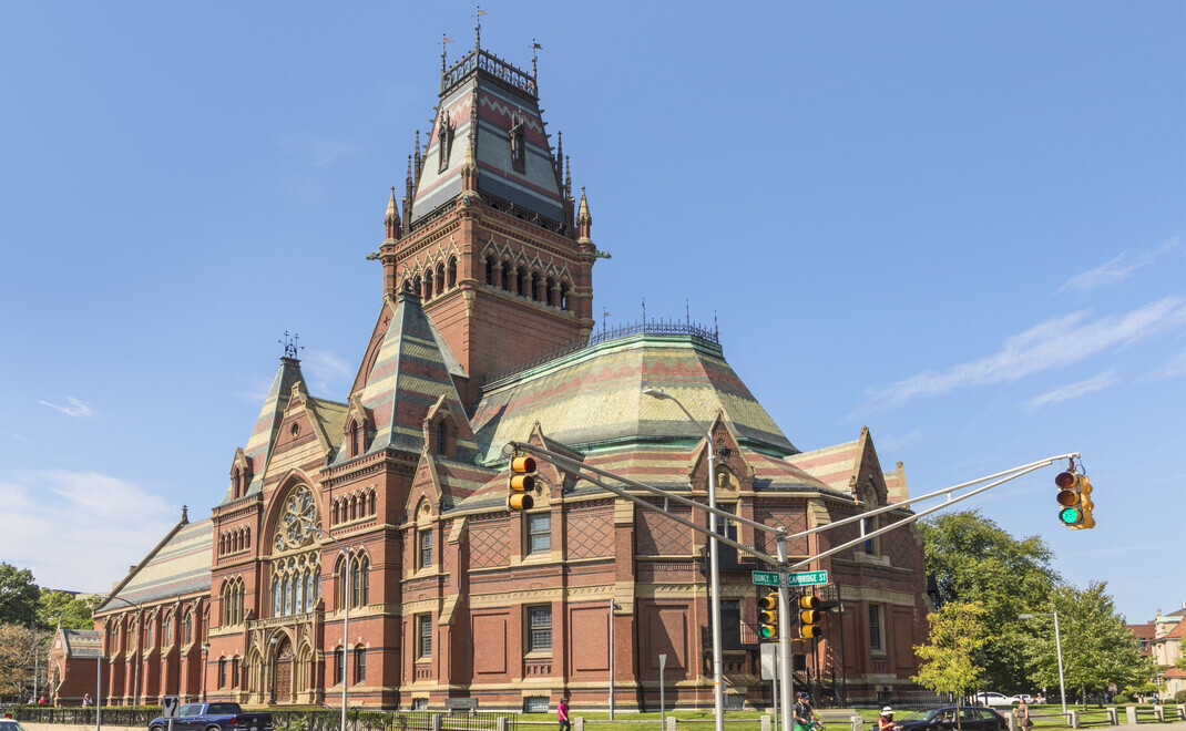 Historisches rotes Backsteingebäude der Harvard Memorial Hall im viktorianischen Gothic-Stil mit hohem Turm, Kupferdach und Spitzbögen, aufgenommen bei blauem Himmel an einer Straßenkreuzung in Cambridge