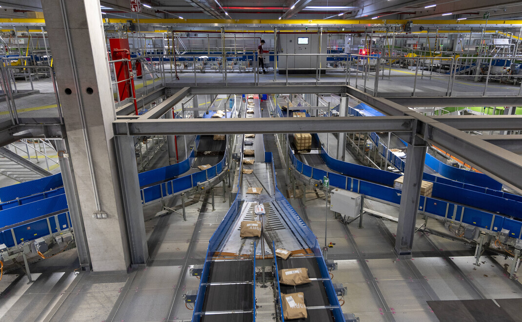 Blick auf ein modernes Logistikzentrum mit mehreren parallelen Förderbändern auf zwei Ebenen. Die Anlage ist in industriellem Design mit blauen Förderbändern, grauen Metallkonstruktionen und gelben Sicherheitsmarkierungen ausgestattet. Auf den Förderbände