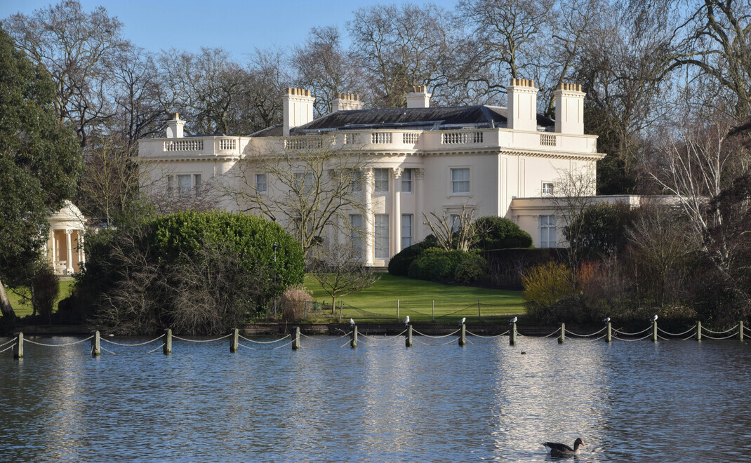 Weiße klassizistische Villa mit hohen Säulen und mehreren Schornsteinen liegt erhöht auf einem gepflegten Grundstück am See im Regent's Park. Das zweistöckige Gebäude mit symmetrischer Fassade ist von Bäumen umgeben. Im Vordergrund ist der See mit einer K